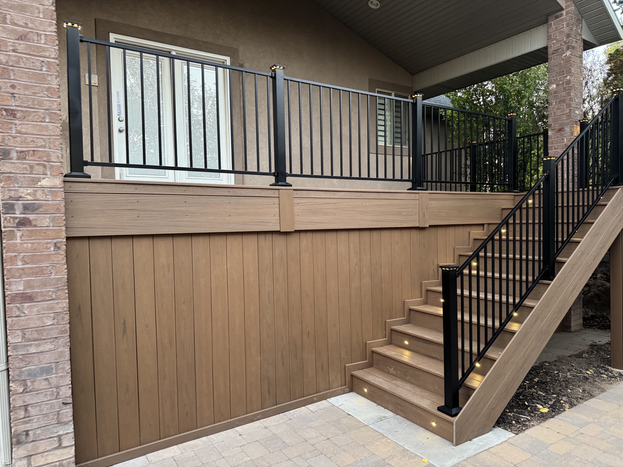Composite deck with black railing and lit stairs beside a brick home