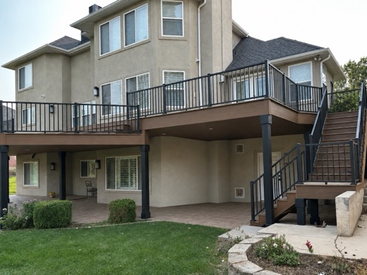 Elevated brown deck wrapping stucco home, black metal and glass railing, stairs to patio
