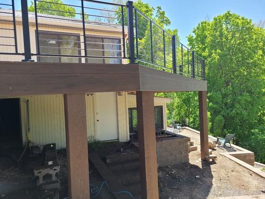 Elevated deck with black horizontal rod railing and brown posts above shaded patio