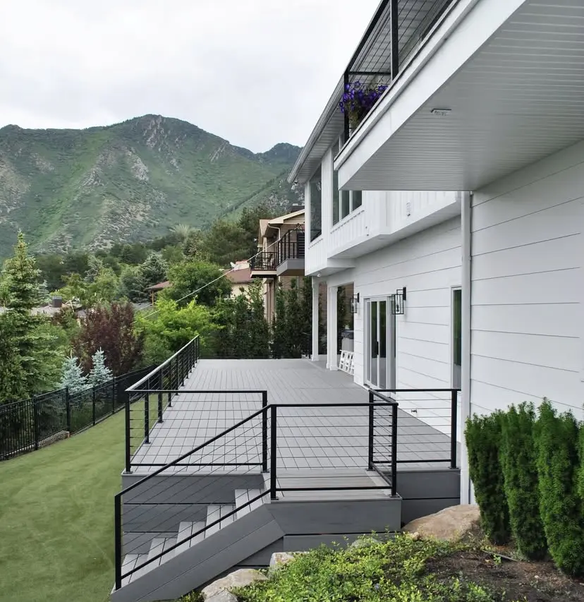 A large porch overlooking a mountainous landscape, with a grey railing, wicker furniture, and potted plants.