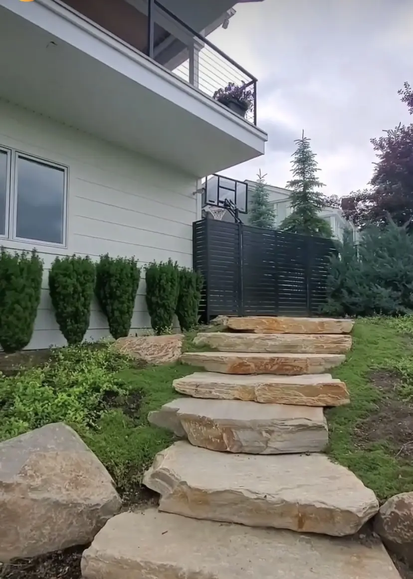 The image shows a gray wooden deck or patio with a railing. The deck leads up to a large building with siding and windows. In the foreground, there is a stone staircase leading up to the deck, and there are tall green trees and shrubs surrounding the building.