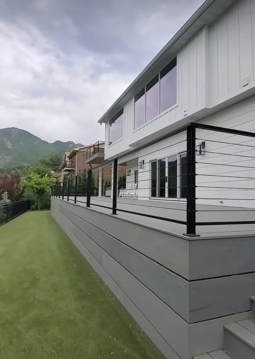The image shows a gray wooden deck or patio with a railing. The deck leads up to a large building with siding and windows. In the background, there are tall green trees and mountains visible against a cloudy sky.