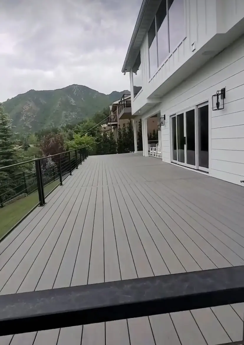 The image shows a gray wooden deck or patio with a railing. The deck leads up to a large building with siding and windows. In the background, there are tall green trees and mountains visible against a cloudy sky.