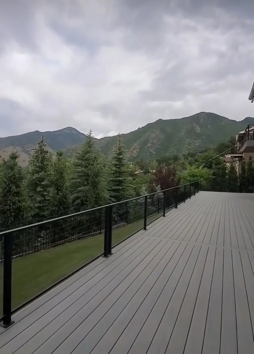 The image shows a large wooden deck or patio with a railing. In the background, there are tall green trees and mountains visible against a cloudy sky.