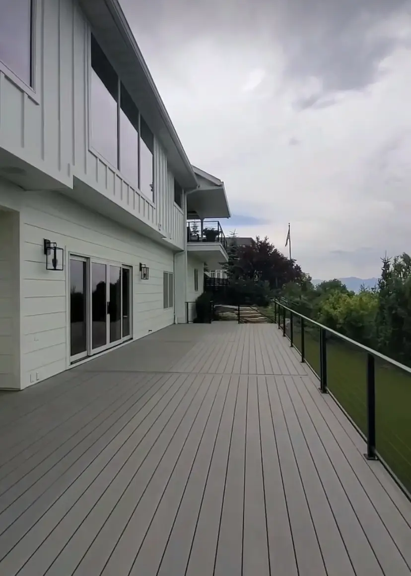 The image shows a gray wooden deck or patio with a railing and mountains visible in the background. The deck leads up to a large building with siding and windows.