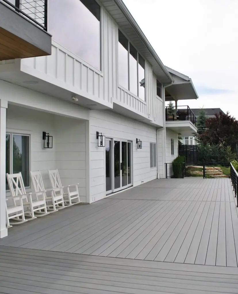 A side view of a modern, multi-story home with a wraparound porch, featuring a gray deck, white siding, and black metal railings.