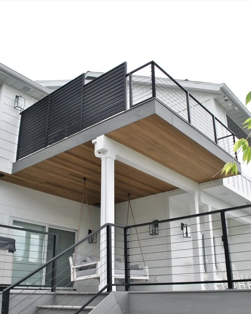 A modern, two-story home with a covered porch and a balcony above, featuring a combination of white siding, wood accents, and black metal railings.