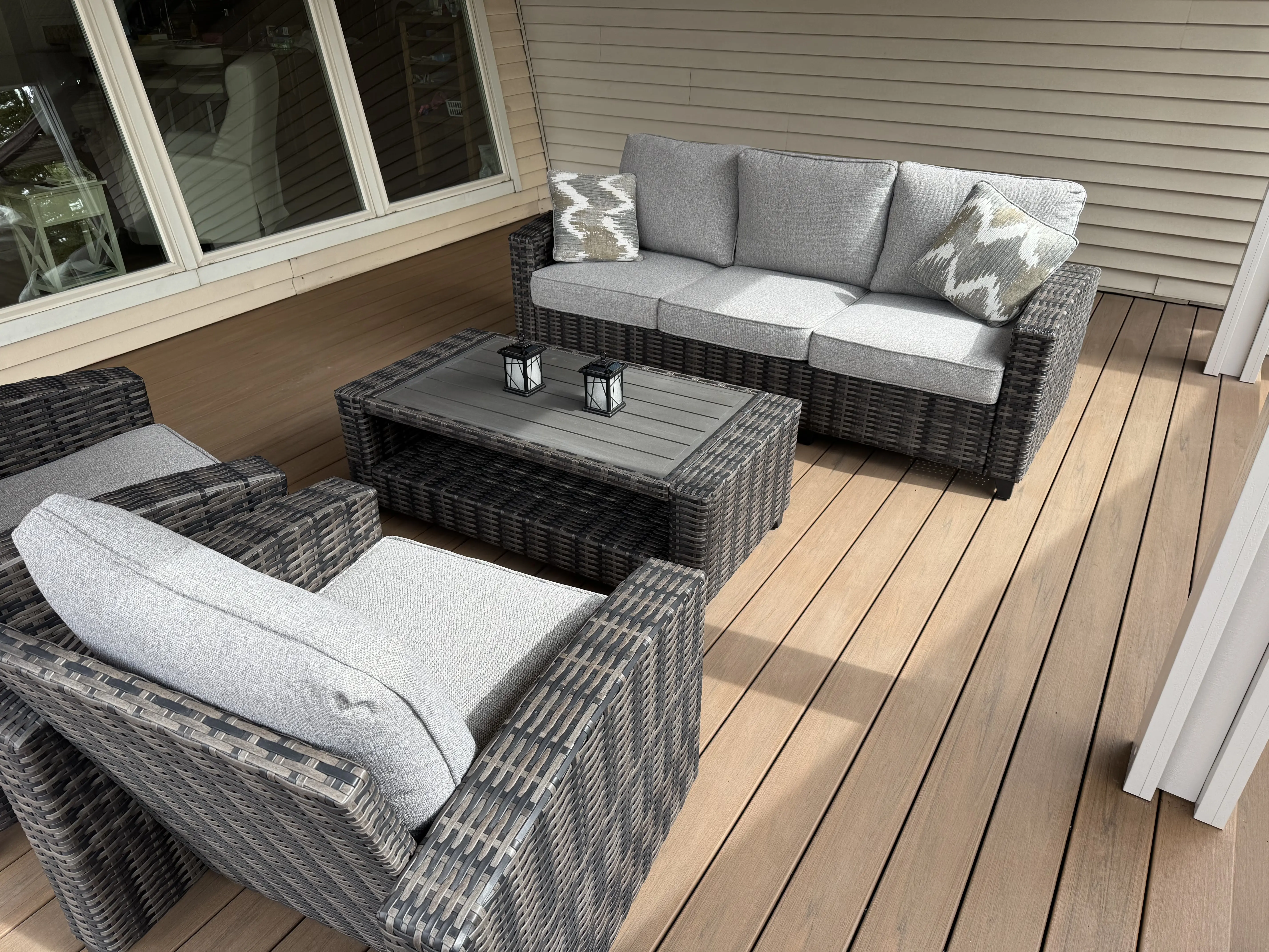 A patio area with comfortable wicker furniture, including a sofa and chairs, set on a wooden deck. There is a low coffee table on the deck, and the exterior of the home is visible in the background.
