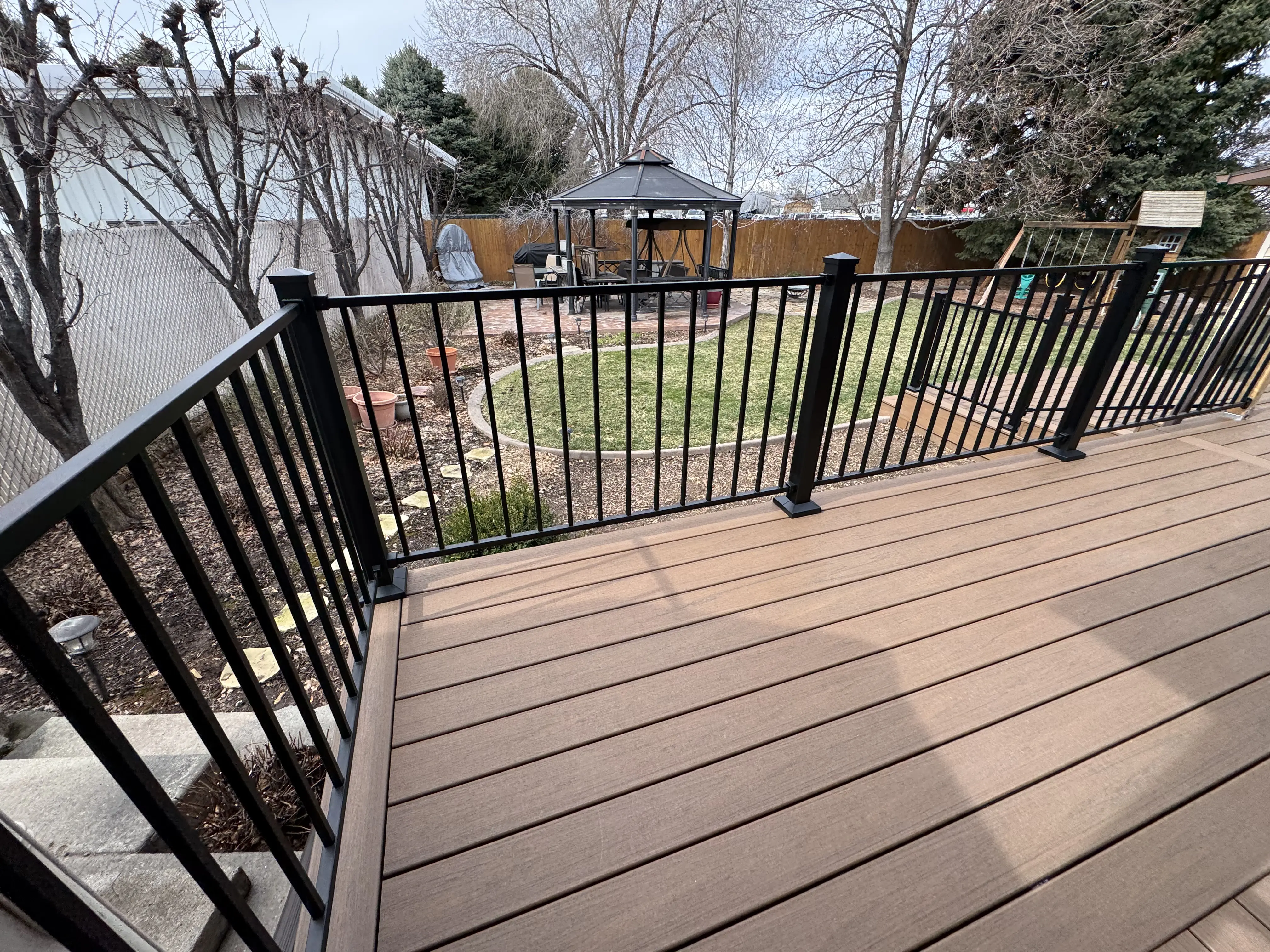 A large wooden deck with a black metal railing, overlooking a yard with a gazebo and trees.