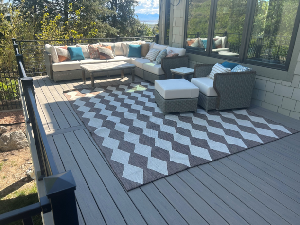 An outdoor patio or deck with a sectional sofa, chairs, and a coffee table. The patio has a chevron-patterned rug and is surrounded by lush greenery, including trees and shrubs.
