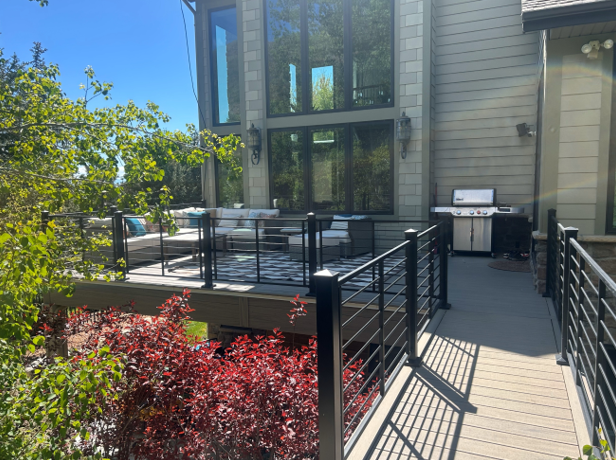 A deck or patio with a wooden railing and steps leading down to the ground. The deck is surrounded by lush greenery, including trees and bushes. The exterior of the home is visible in the background.
