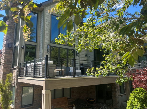 A deck or patio with a wooden railing, surrounded by trees and greenery. The deck leads to the exterior of a home, which has large windows and a covered porch area.