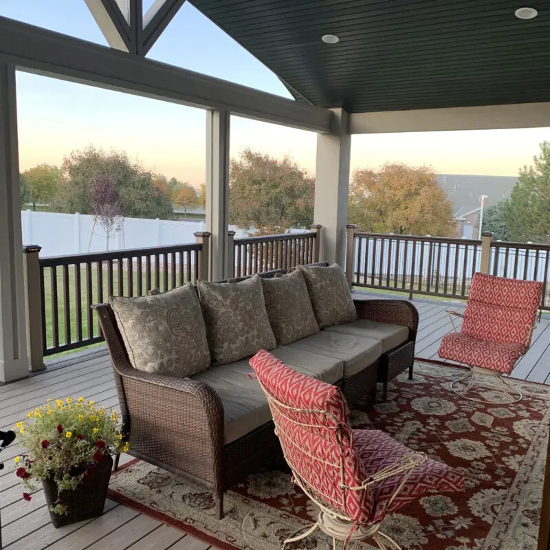 A covered porch with a gray wood deck, wicker furniture, and a decorative rug, overlooking a backyard with trees.