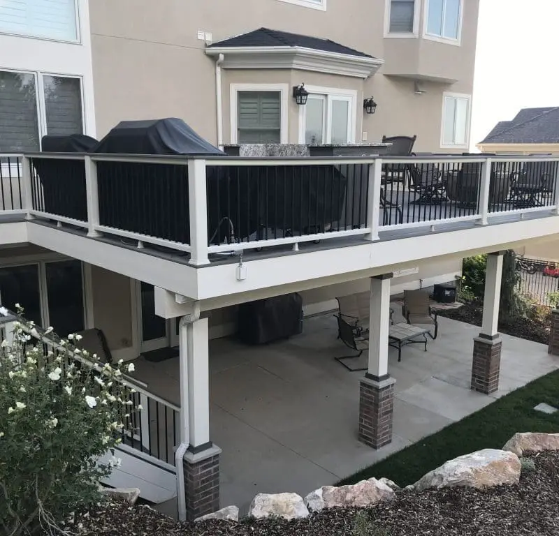 Two-level white deck with black metal railings, covered patio beneath, brick support columns, and manicured lawn with landscaping