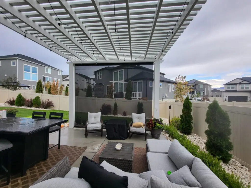 backyard patio space that has a white pergola and drop down privacy screen/sun shade. Outdoor furniture, dining furniture and decor are under the pergola.