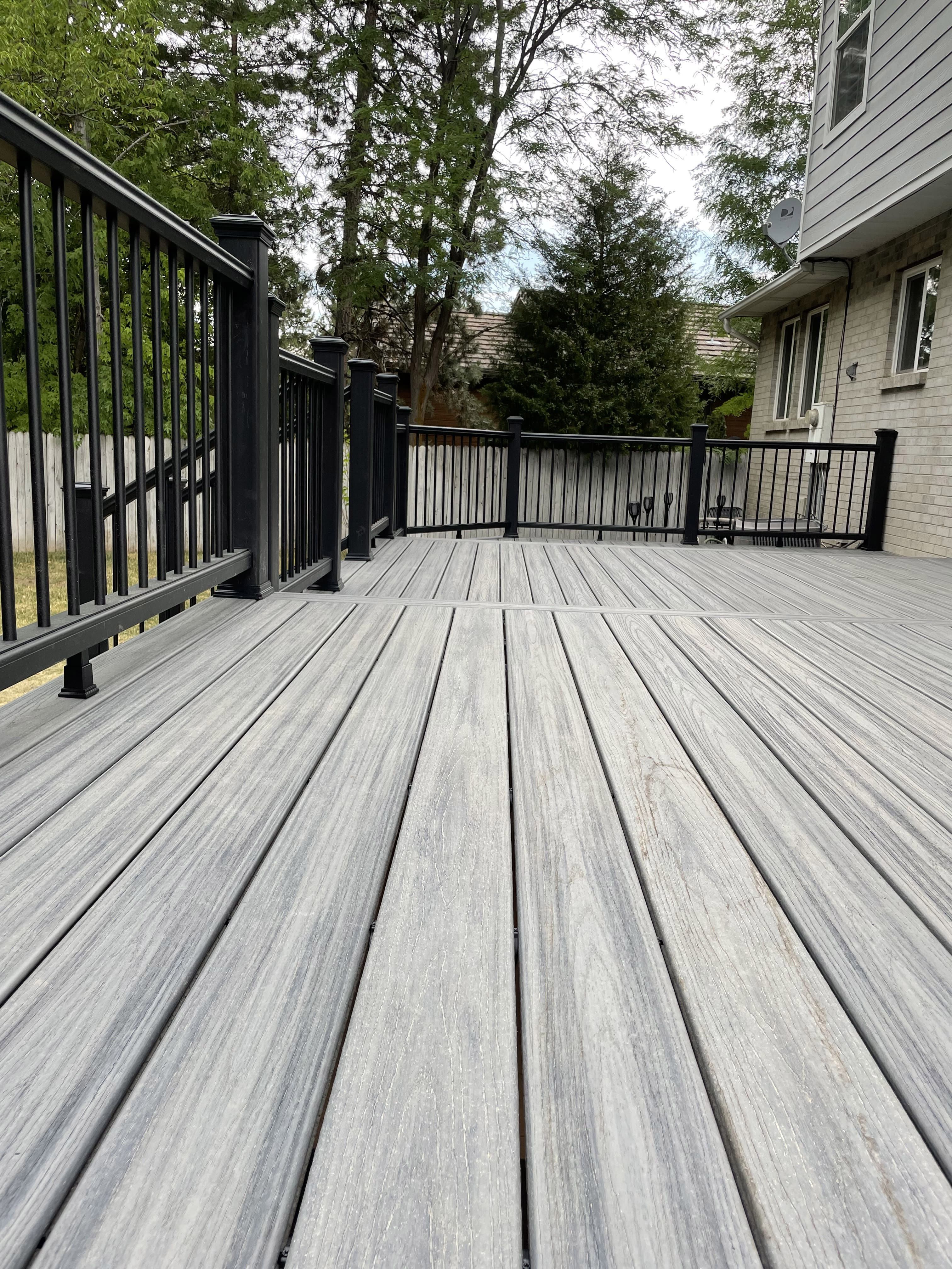 A gray wooden deck with a metal railing. The deck is attached to a residential building and is surrounded by trees.