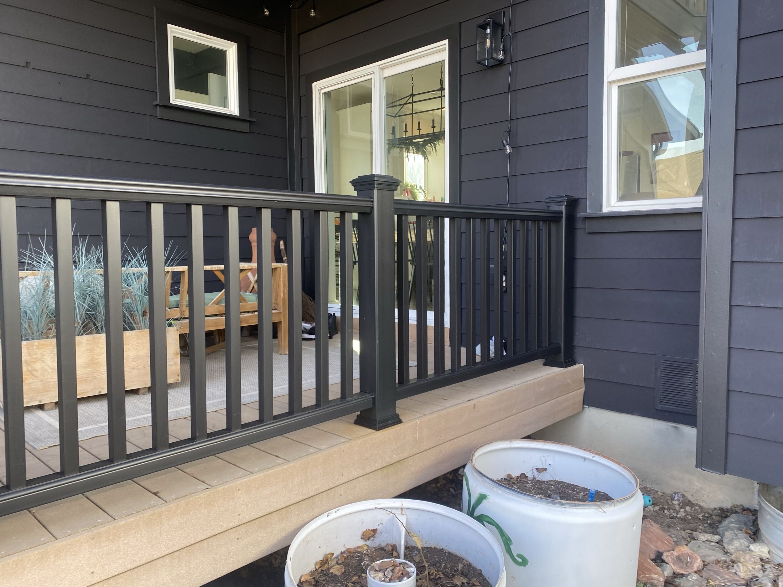 A wooden deck with a black railing overlooking a yard with plants and trees. There are two white containers with soil on the deck.