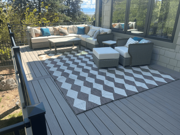 An outdoor patio or composite deck with a sectional sofa, chairs, and a coffee table. The patio has a chevron-patterned rug and is surrounded by lush greenery, including trees and shrubs.