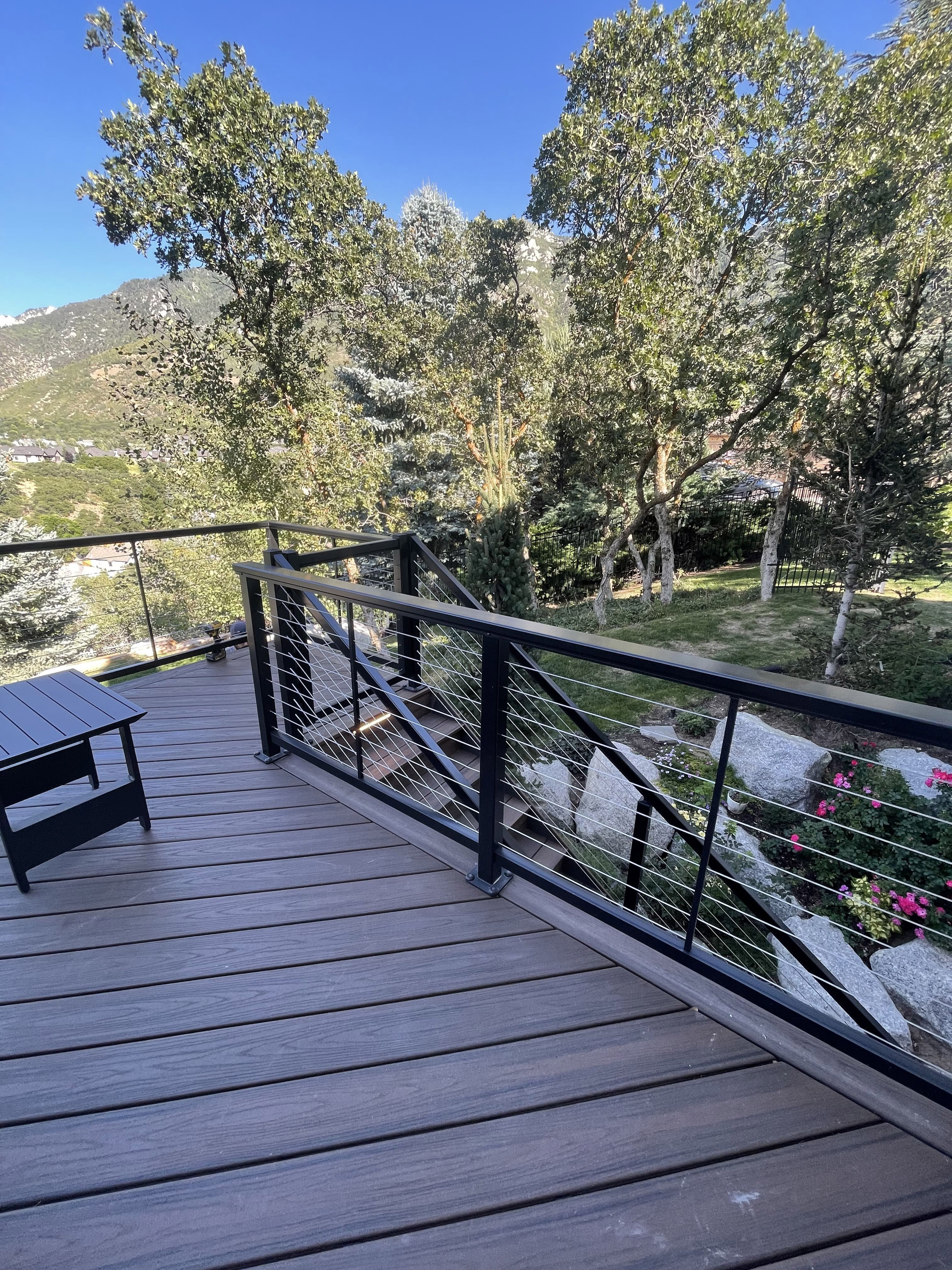 A wooden deck with a metal railing overlooking a snowy mountain landscape. The deck is surrounded by lush greenery and there is a small table with chairs on the deck.