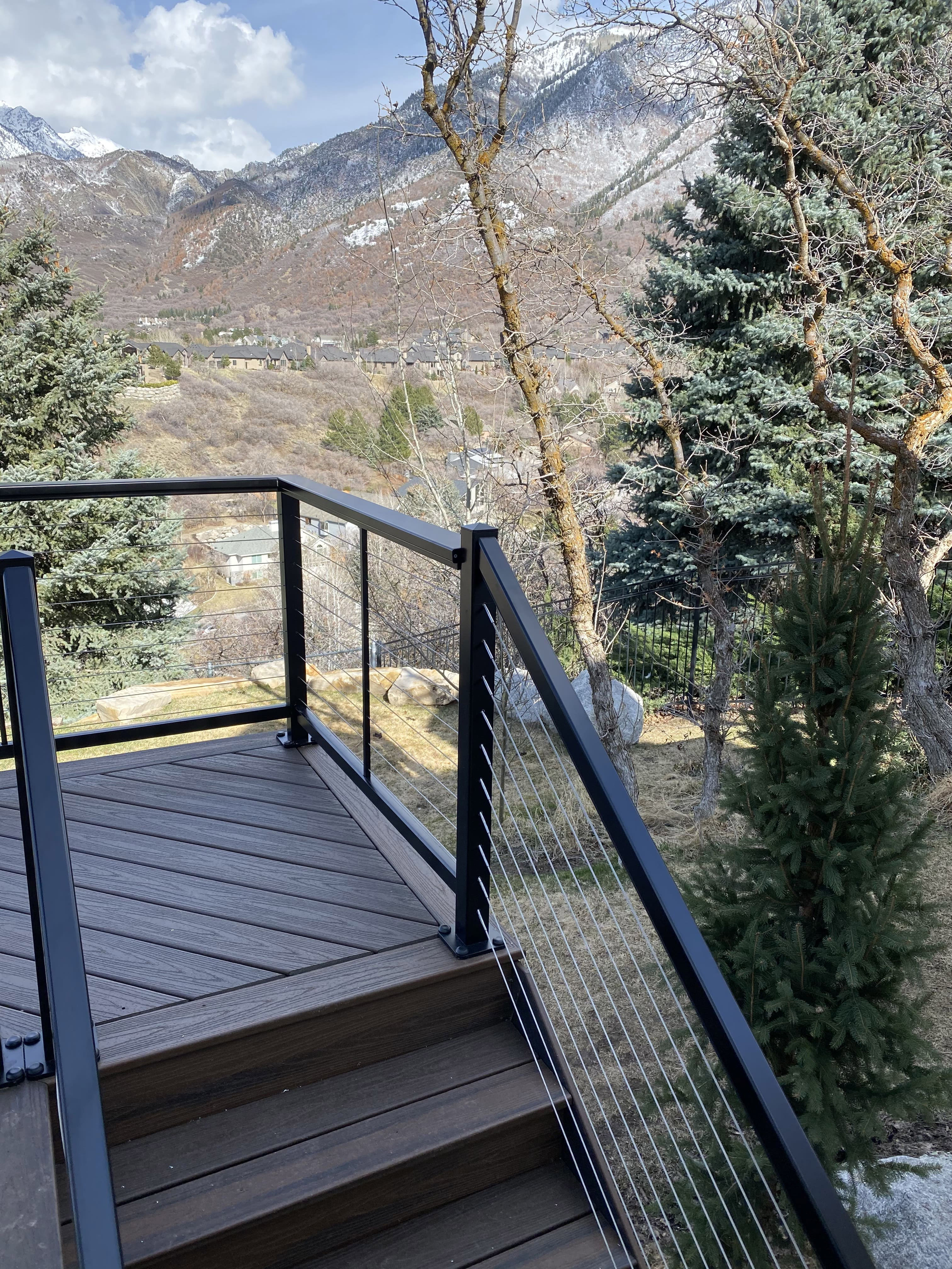 A wooden deck with a metal railing overlooking a snowy mountain landscape. The deck is surrounded by pine trees and has a cozy outdoor seating area.