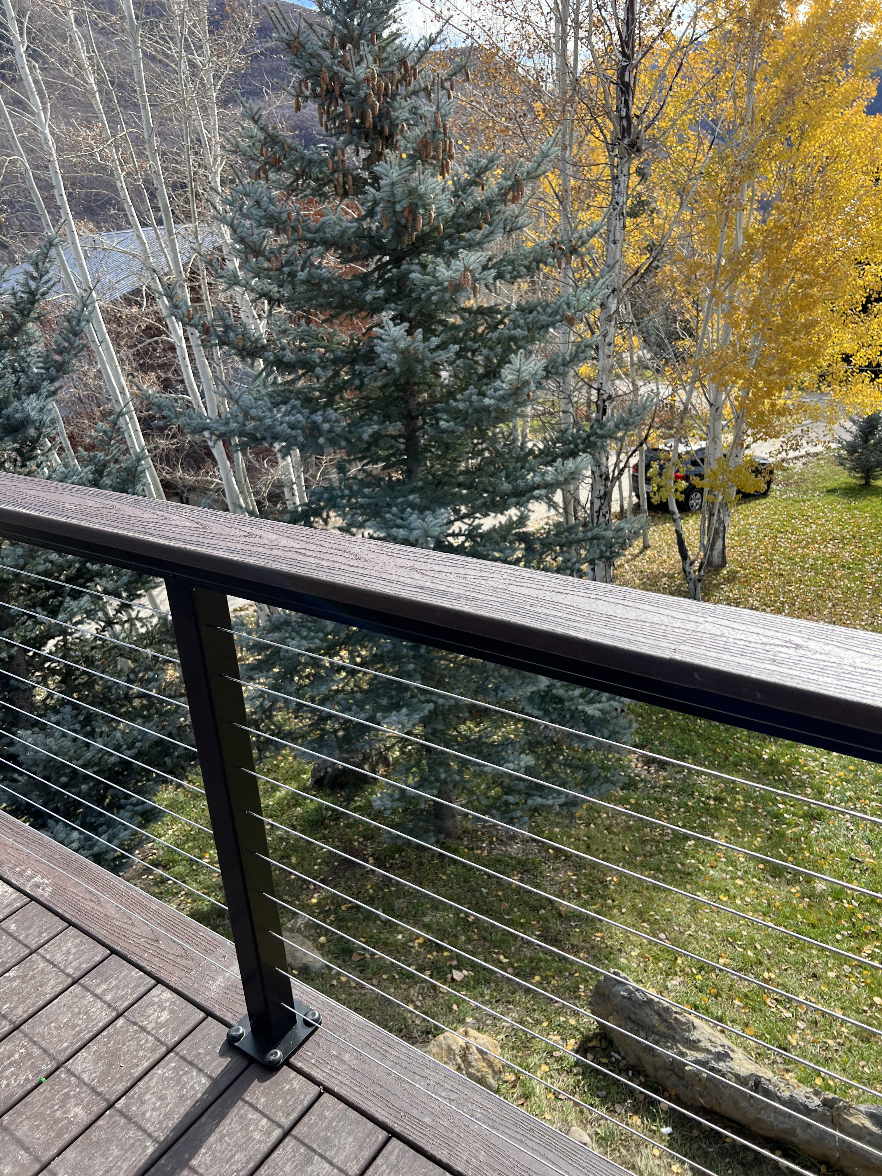 A wooden deck with a metal railing overlooking a snowy mountain landscape. The deck is surrounded by trees with yellow and green foliage.