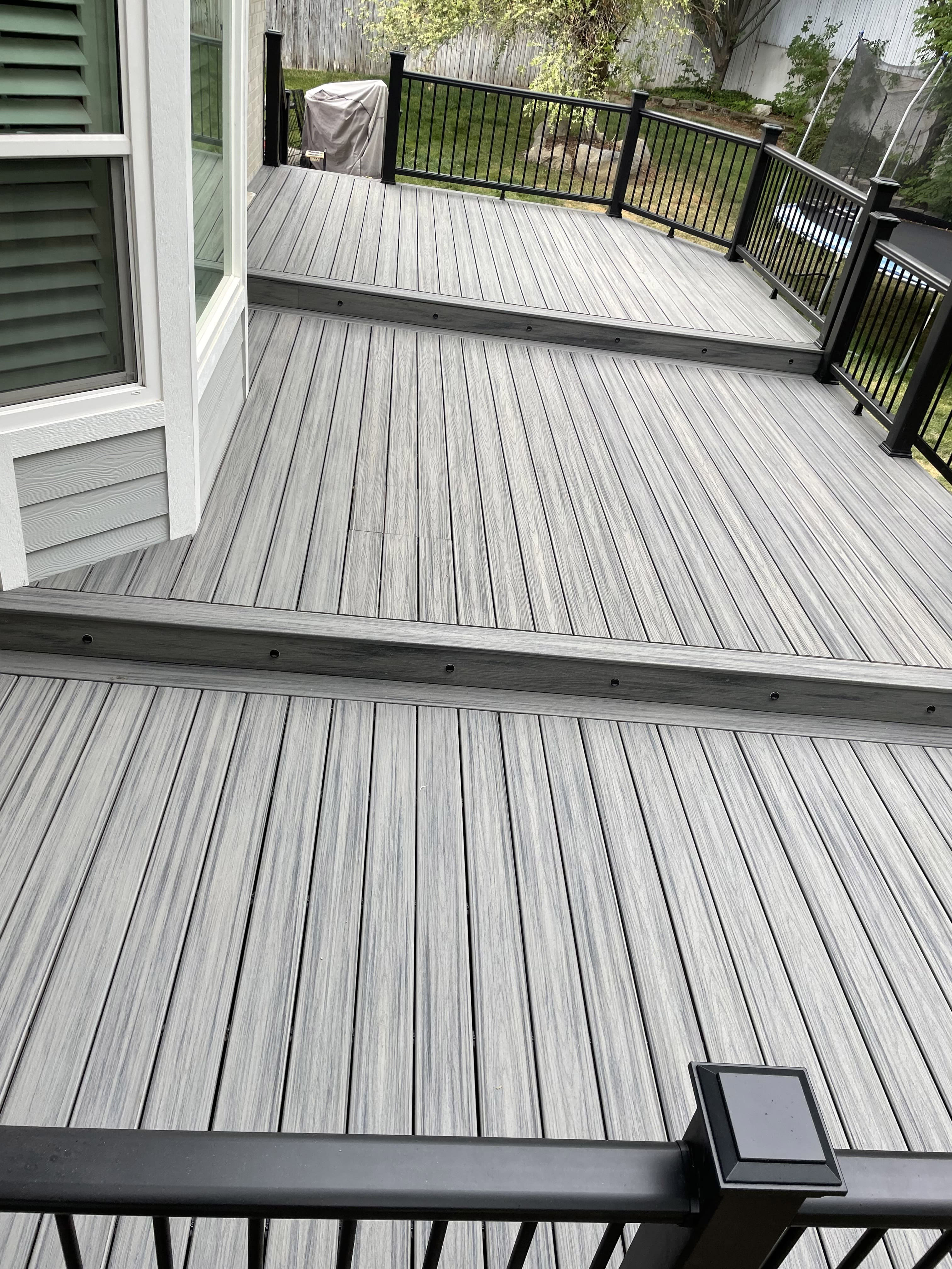 Trex-Island-Mist-0-28 A large gray wood deck with a black metal railing overlooking a wooded backyard.
