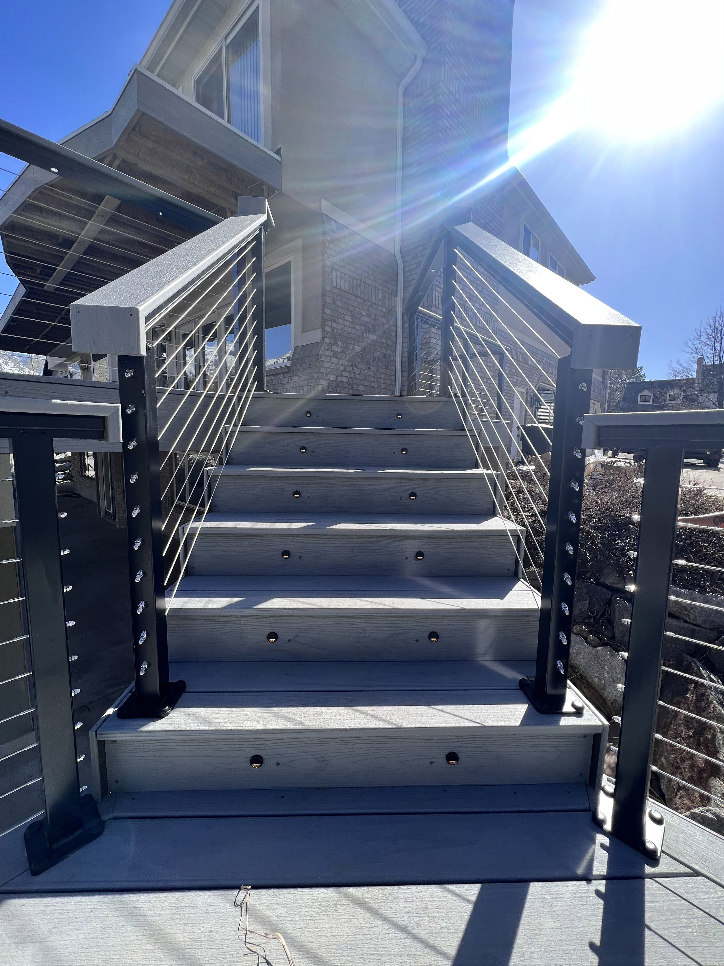 Trex-Lineage-Rainier-Decking-13-37 A set of concrete steps with a metal handrail leading up to a building entrance, with the sun shining brightly.