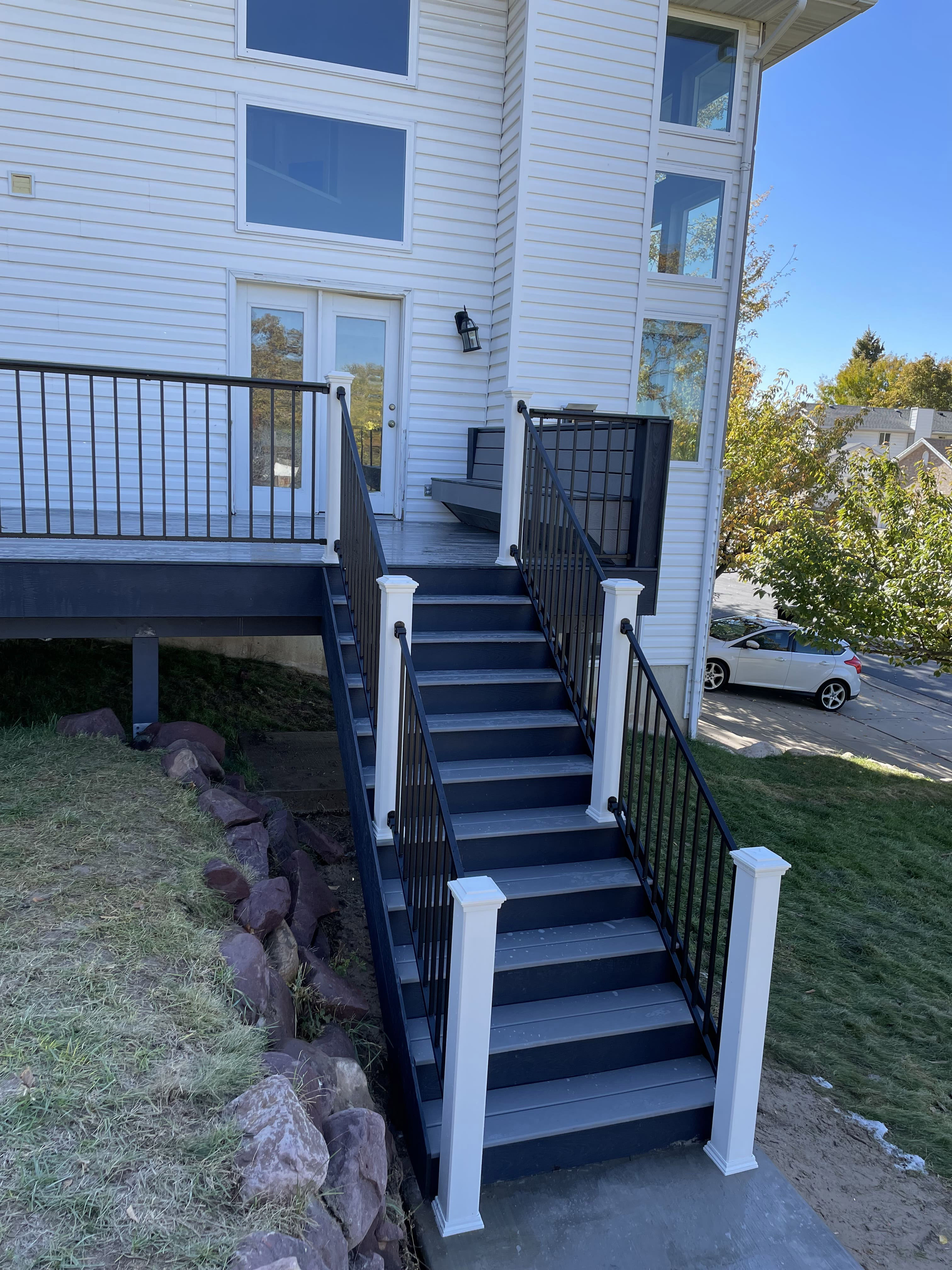 Trex-Pebble-Grey-and-winchester-grey-deck-and-stairs-5 A wooden staircase with black railings leading up to a second-floor porch or balcony of a residential building. The staircase has a sturdy, modern design and is surrounded by lush greenery.
