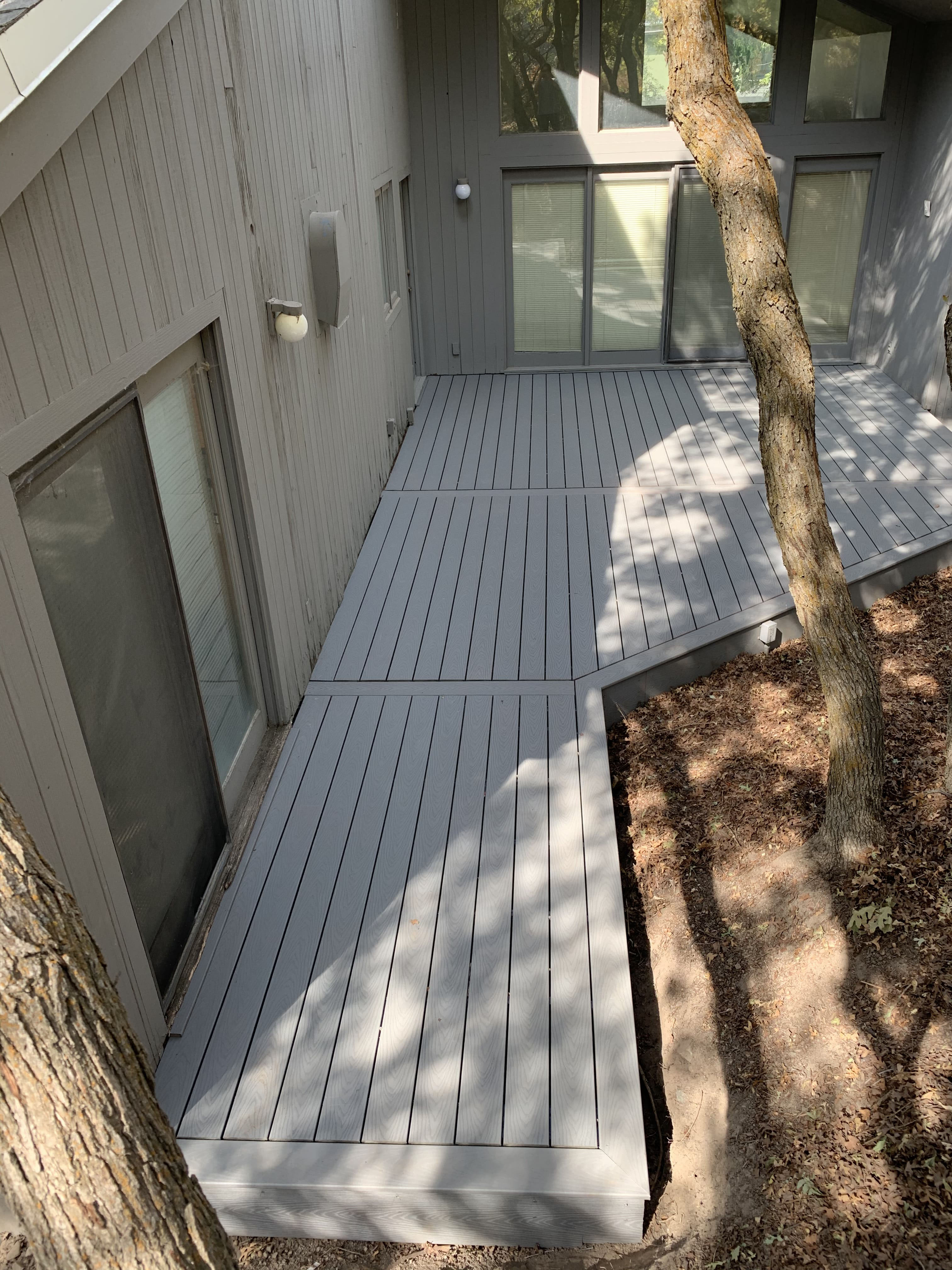 Trex-Select-Pebble-Grey-11-40 A gray wooden deck with a tree trunk and shadows from the overhead branches.
