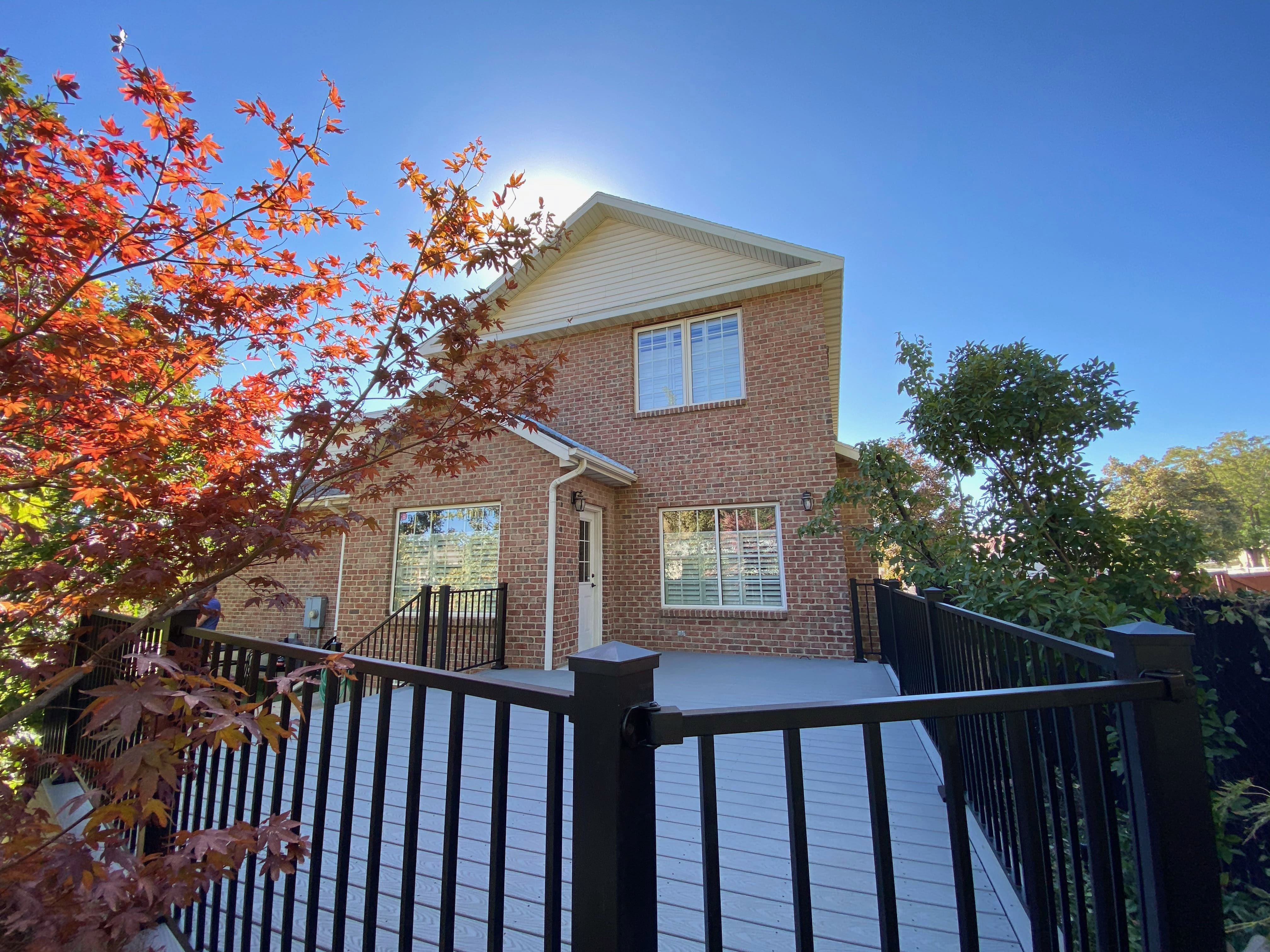 Trex-Select-Pebble-Grey-13-48 A two-story brick house with a porch and trees in the foreground, against a bright blue sky.