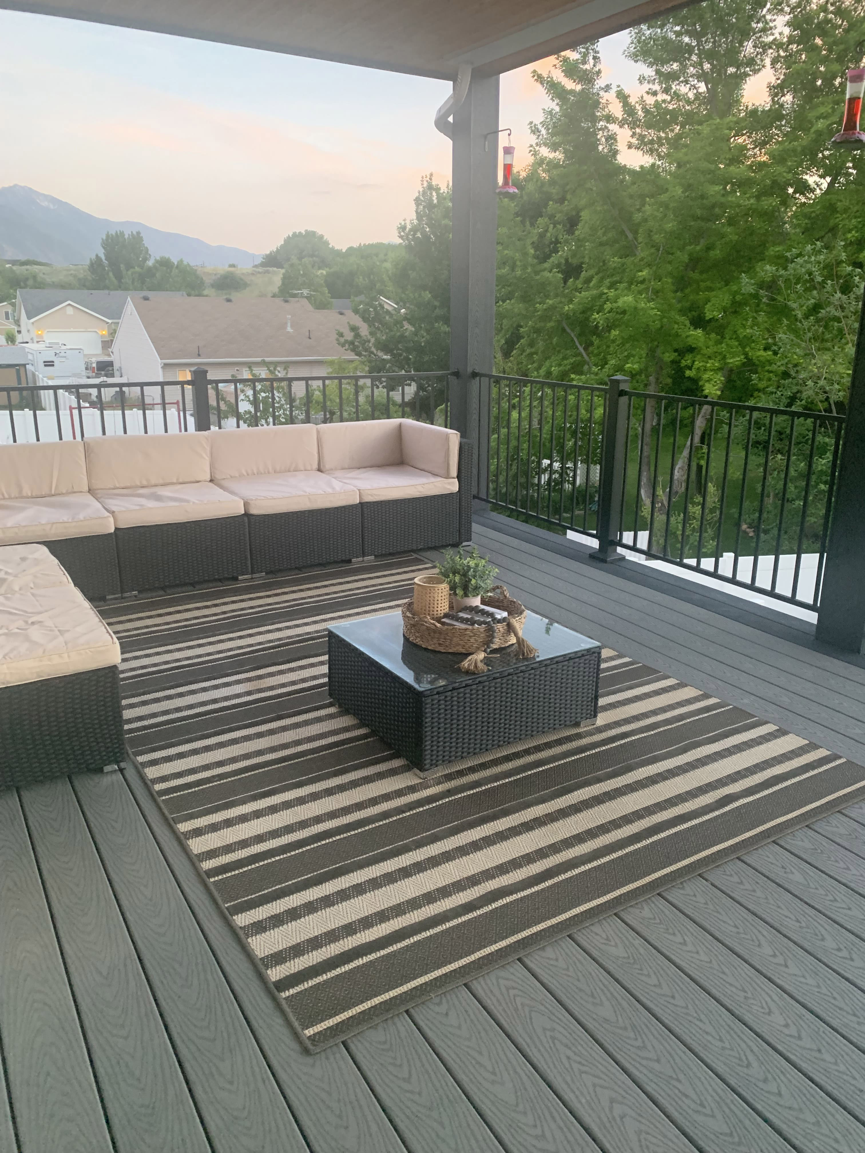 Trex-Select-Pebble-Grey-16 A cozy outdoor living space on a wooden deck, with a sectional sofa and a low, square coffee table. The deck has a striped rug and overlooks a scenic mountain landscape.