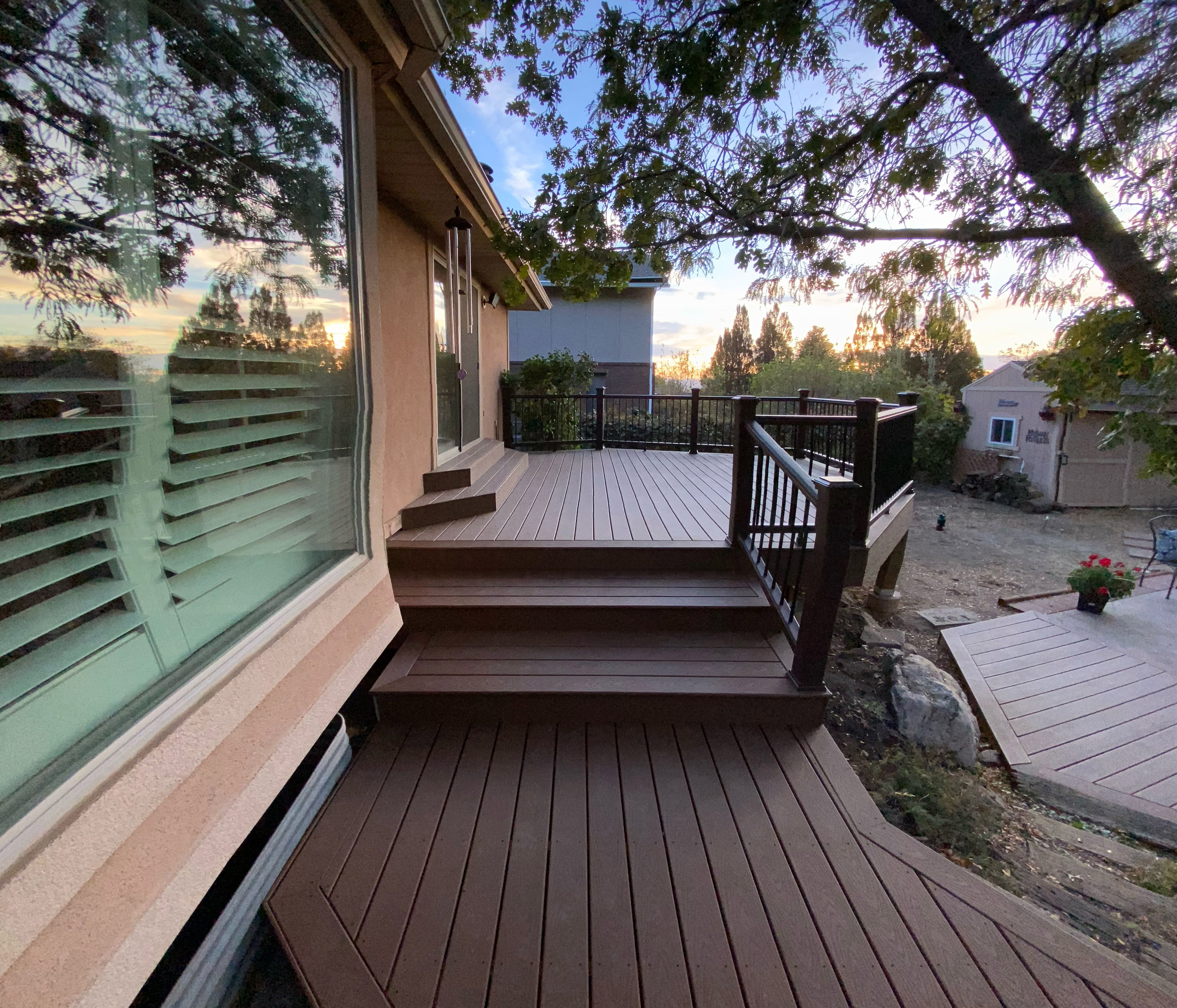 Trex-Select-Saddle-11-63 A wooden deck with steps leading up to it, surrounded by trees and a scenic mountain view in the background. The deck has a black railing and a glass panel on the side.