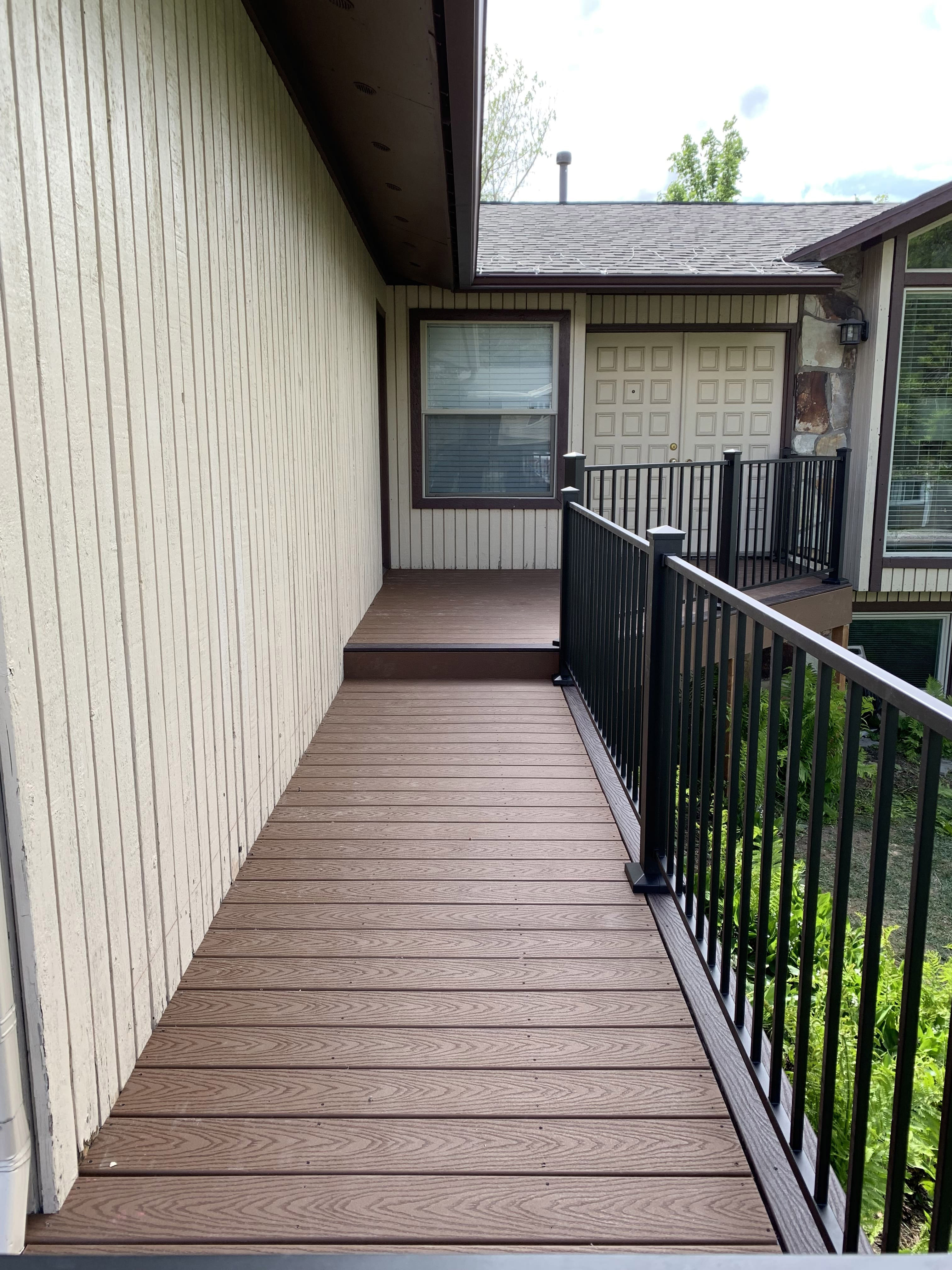 Trex-Select-Saddle-57 A covered porch with a wooden deck and black metal railing. The porch leads to the entrance of a house with siding and has a view of the surrounding trees and landscape.