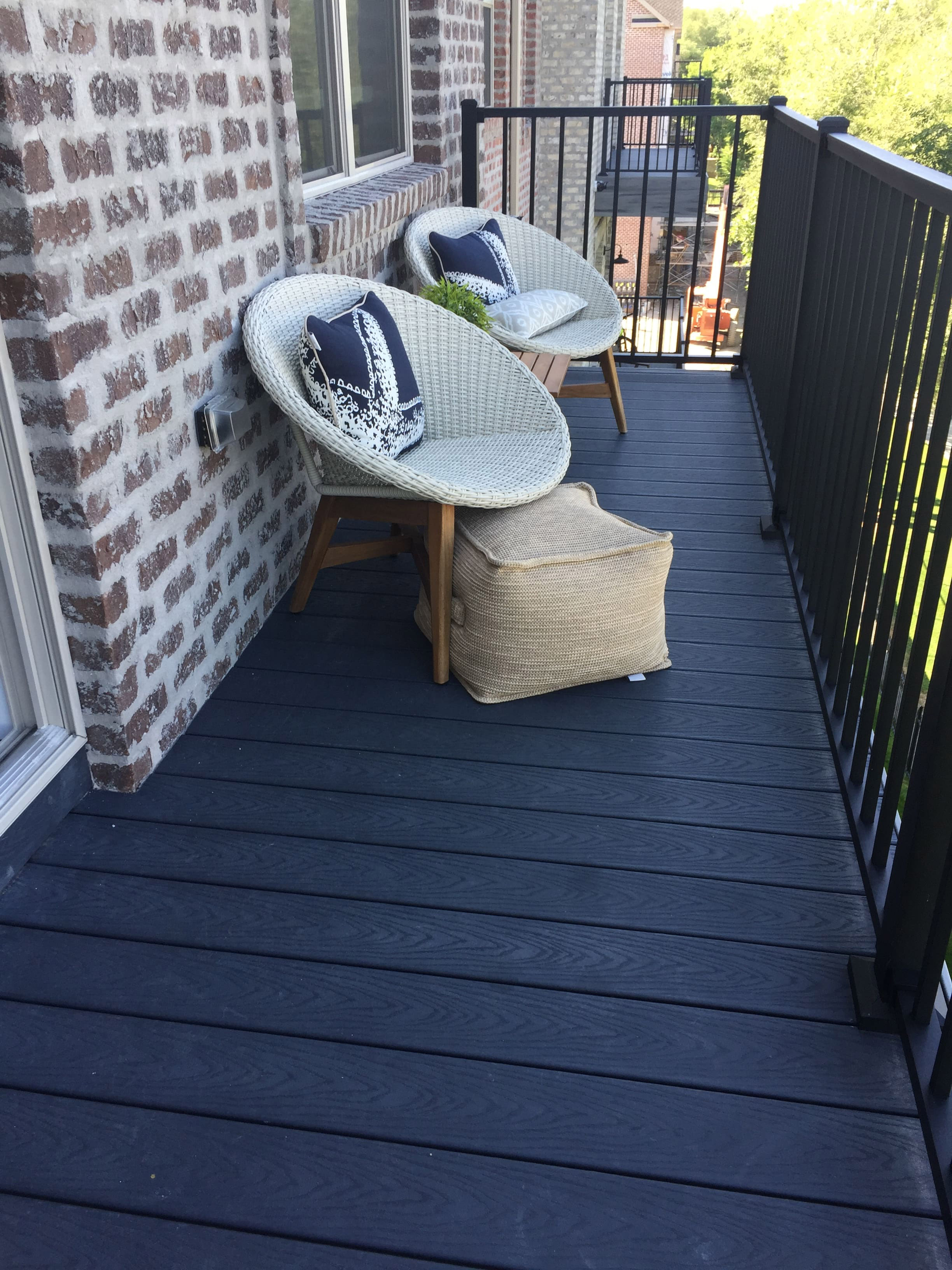 Trex-Select-Winchester-Grey-1-33 A balcony with two wicker chairs and small tables, surrounded by a brick wall and overlooking a tree-lined neighborhood.