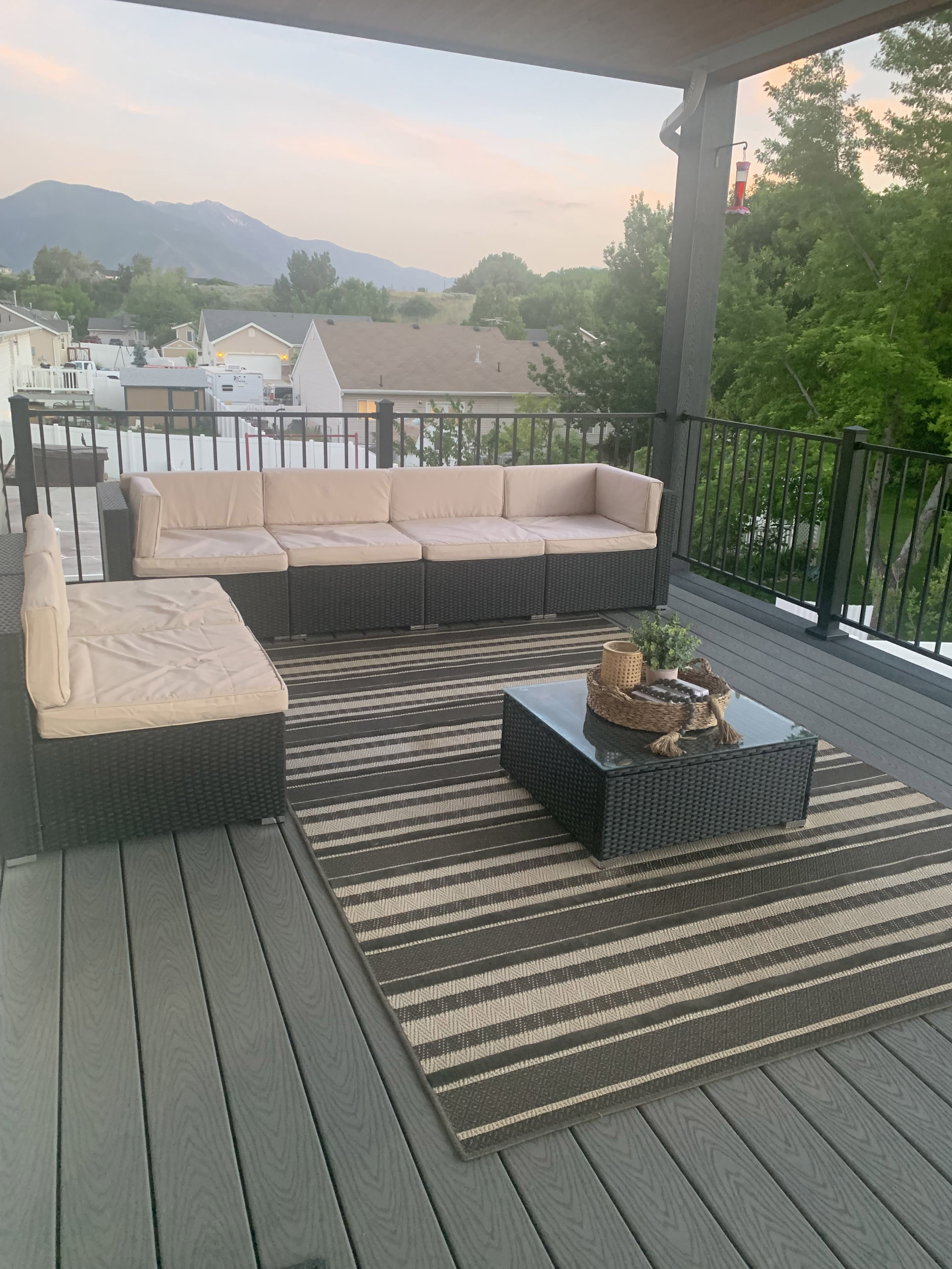 Trex-Select-pebble-grey-and-winchester-grey-3 A patio with a sectional sofa, coffee table, and striped rug, with a mountain view in the distance.