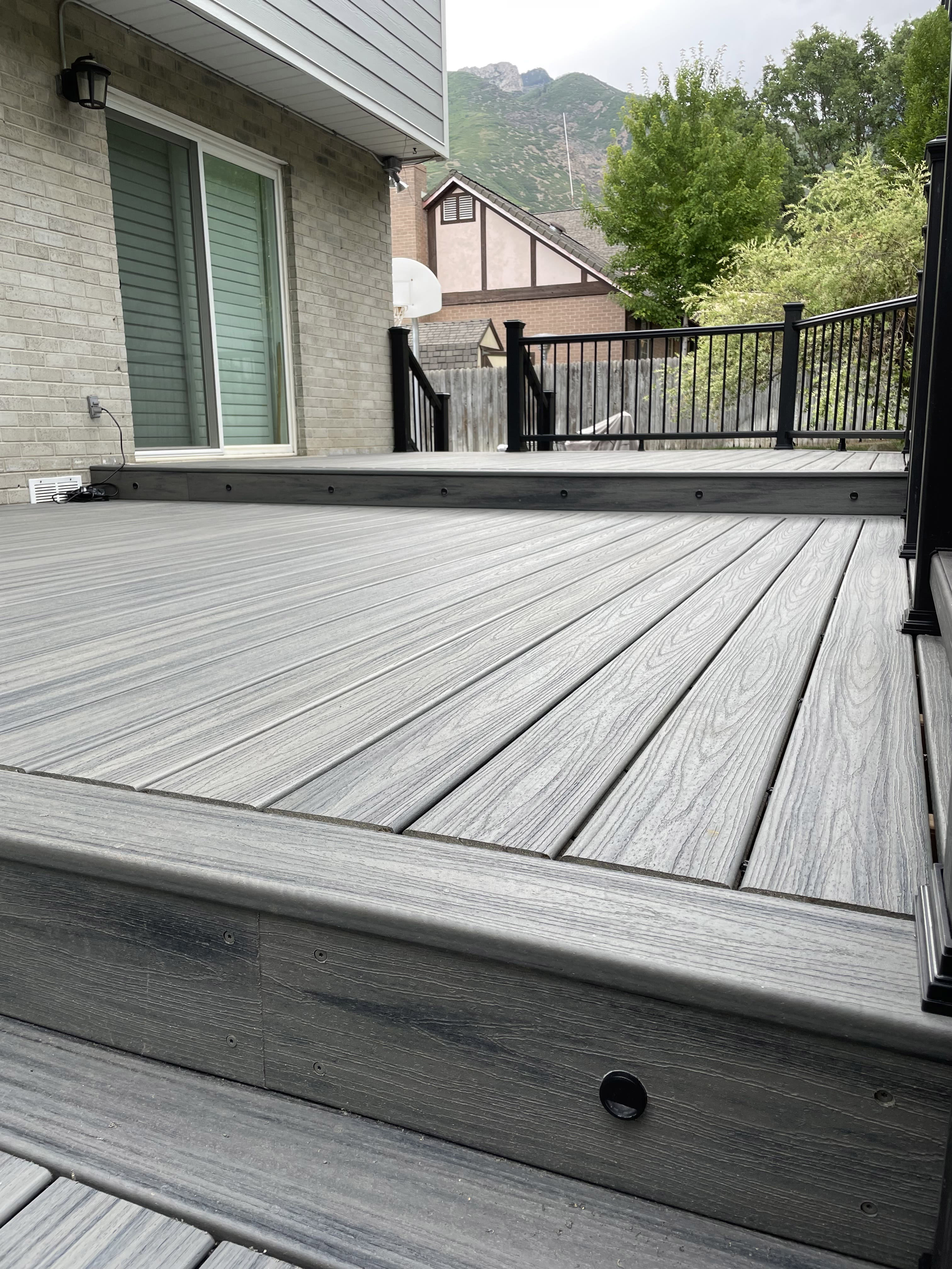 Trex-Transcend-Island-Mist-22 A gray wood deck with a black metal railing, extending out from a house with a stone foundation and surrounded by trees.