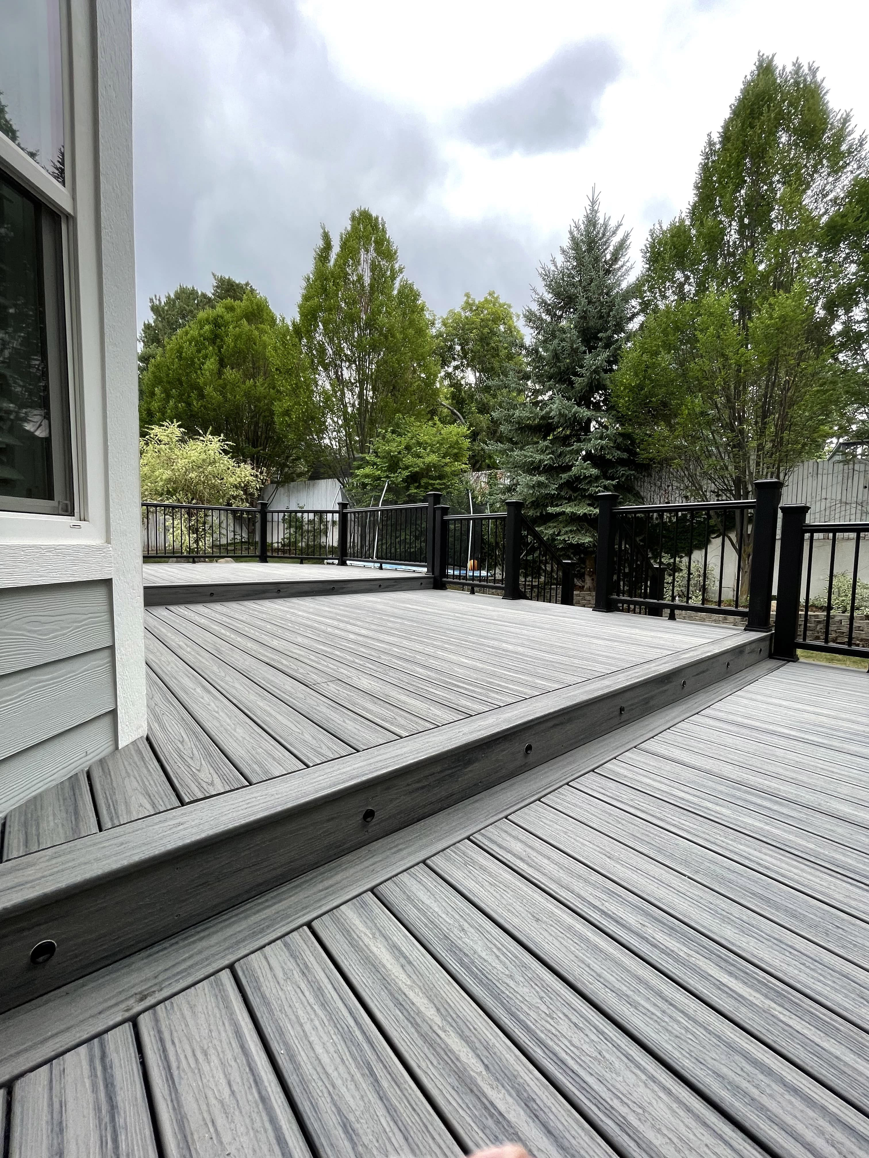 Trex-Transcend-Island-Mist-23-12-19 A wooden deck with a metal railing overlooking a lush green wooded area on a cloudy day.