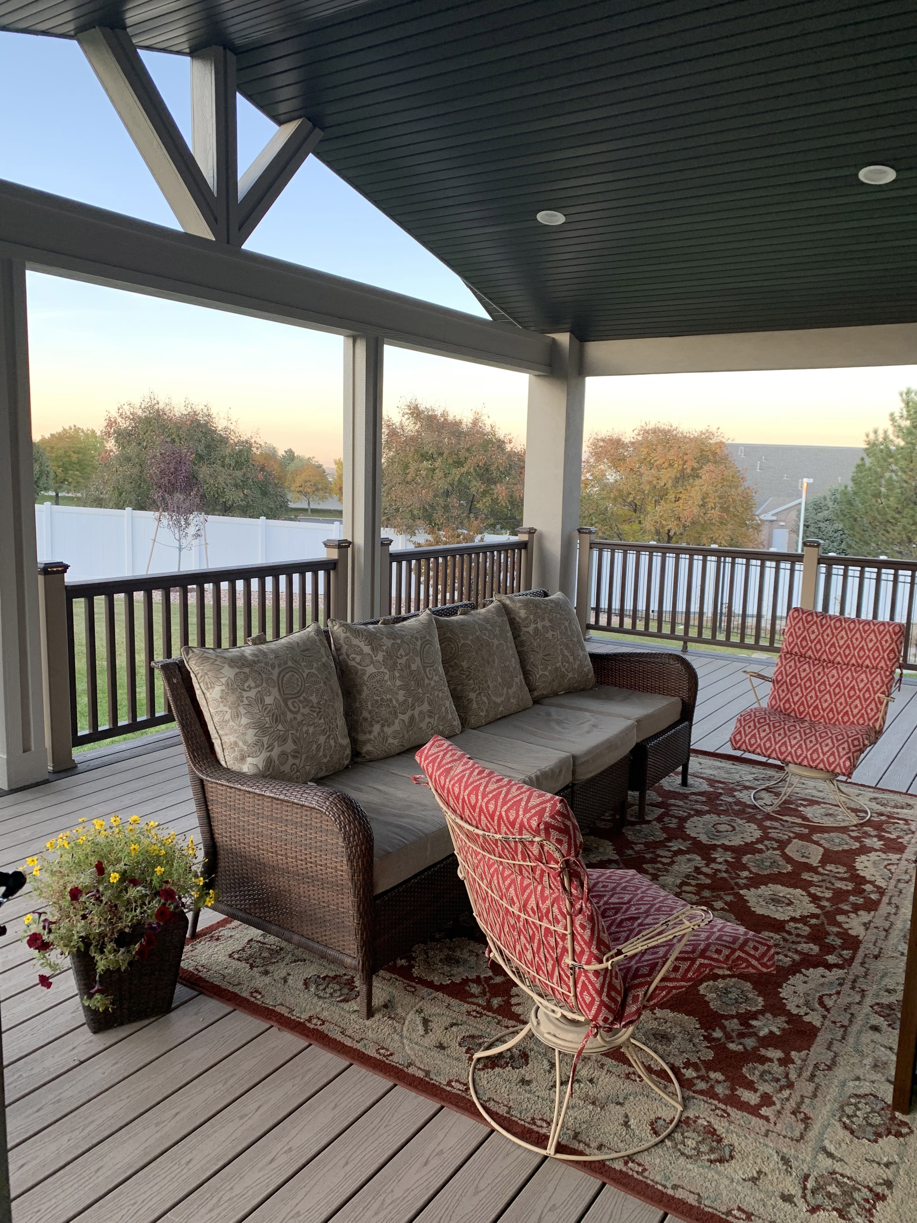 Trex-Transcend-Rope-Swing-with-Vintage-Lantern-21 A covered porch with a gray wood deck, wicker furniture, and a decorative rug, overlooking a backyard with trees.