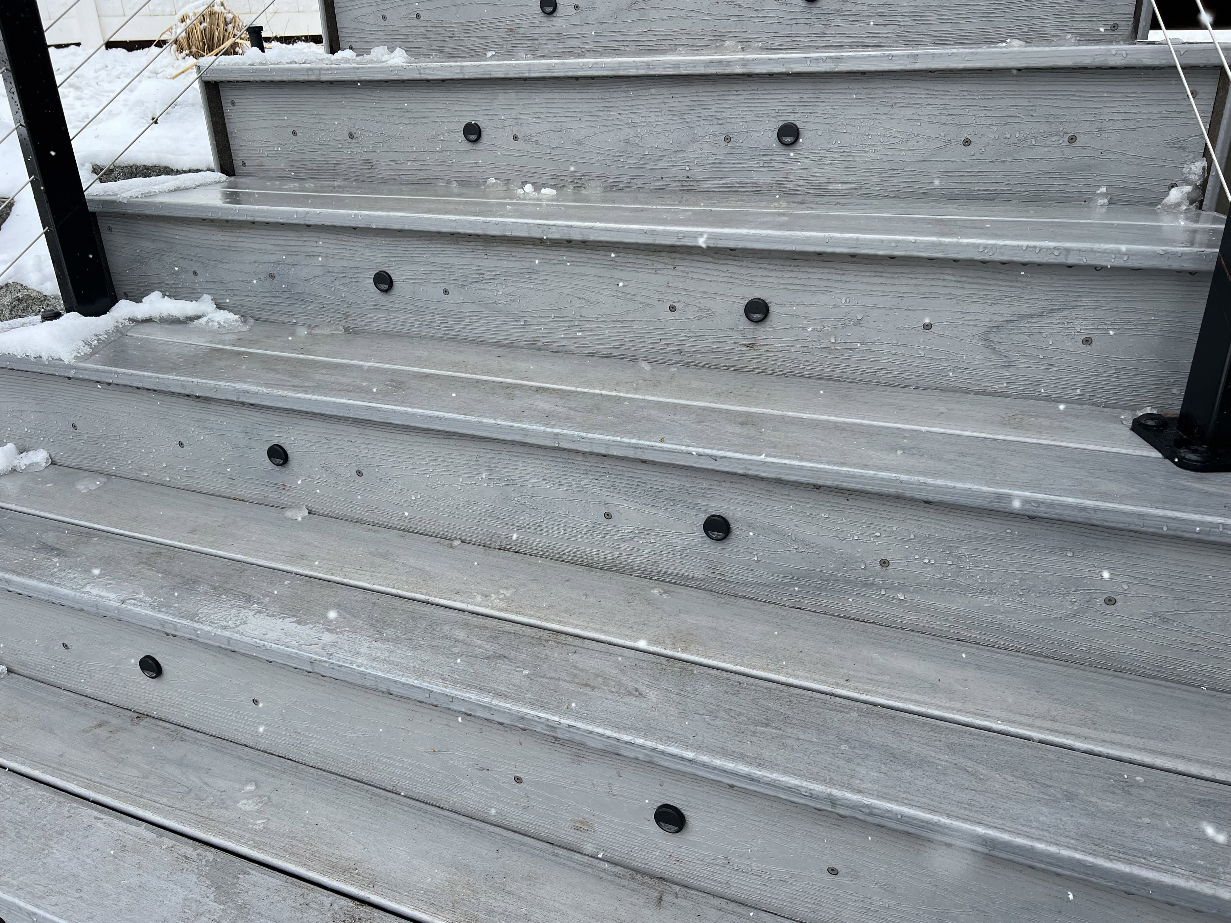 Trex-lineage-biscayne-with-lighting-1-8 A set of snow-covered wooden steps with a metal railing leading up to a building entrance.