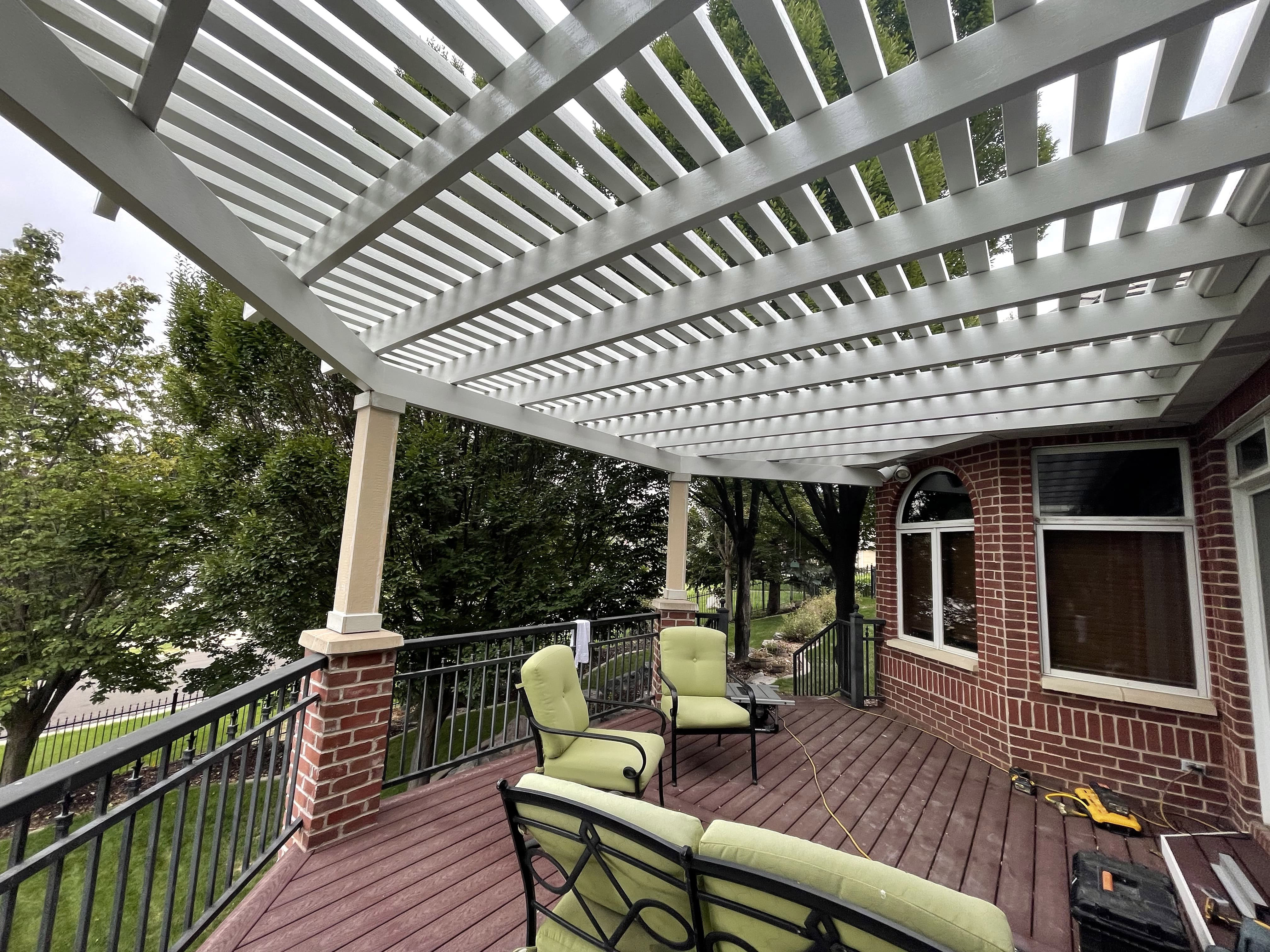 A white, slatted pergola structure with columns supporting the roof, covering an outdoor deck or patio area. The deck has wicker furniture, including chairs and a sofa.